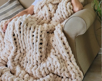 May include: A large, cream-coloured chunky knit blanket is draped over a sofa. The blanket is made of thick, soft yarn and has a textured, woven pattern.