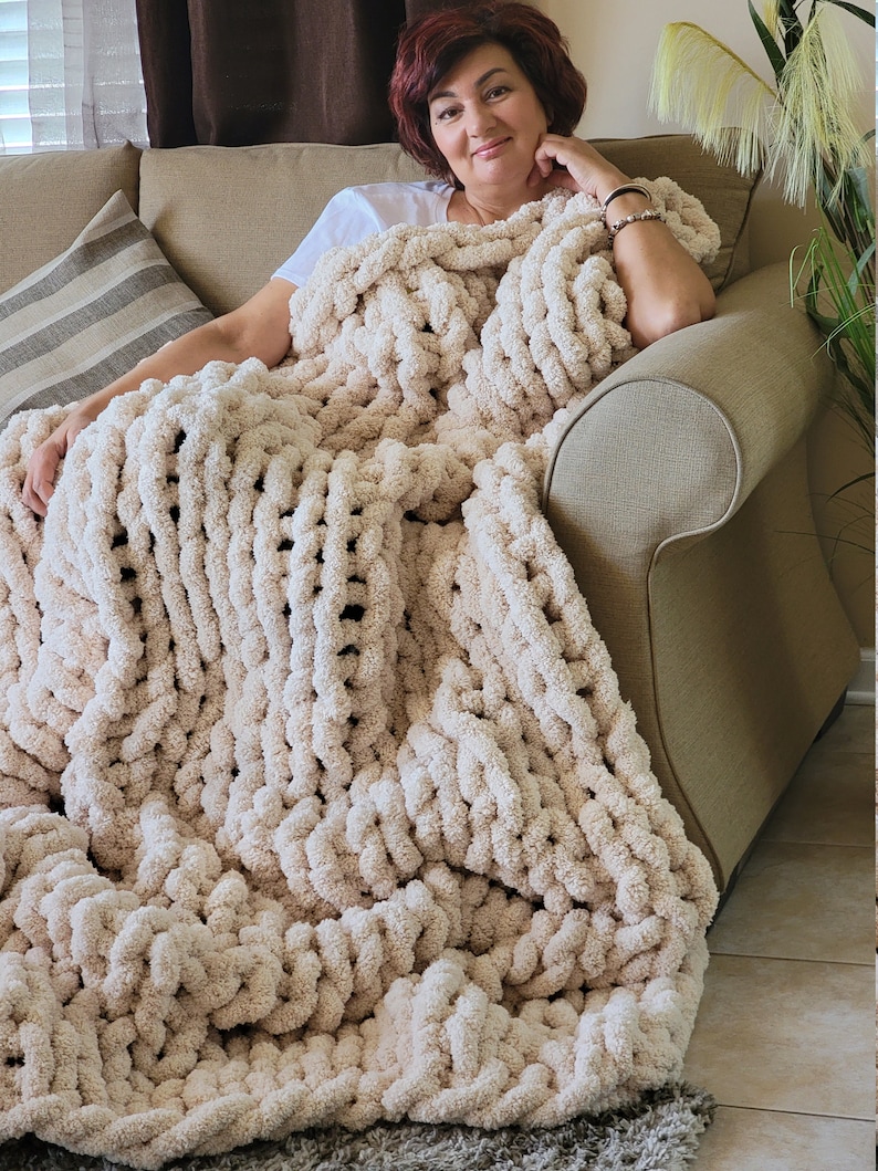 May include: A large, cream-colored chunky knit blanket is draped over a couch. The blanket is made of thick, soft yarn and has a textured, woven pattern.