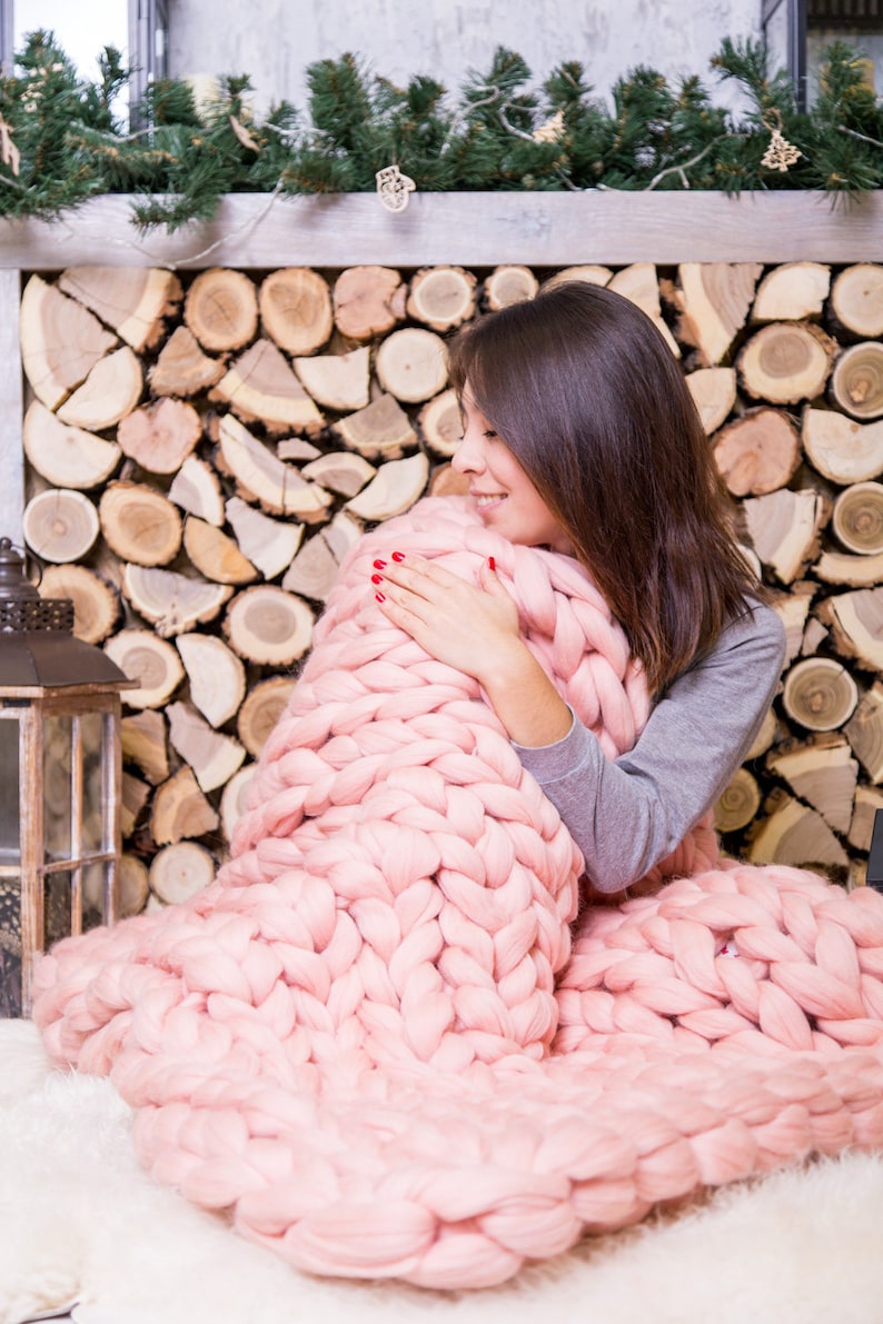 May include: A woman is wrapped in a large, chunky knit pink blanket. The blanket is made of thick yarn and has a soft, cozy texture. The woman is sitting in front of a fireplace with a pile of logs stacked behind her.