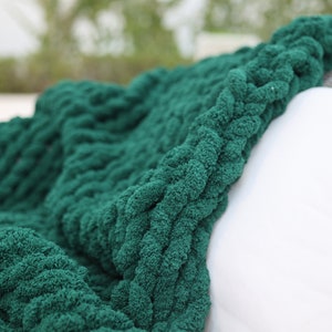 May include: A soft, green knitted throw blanket with a chunky cable knit pattern. The blanket is draped over a white surface.
