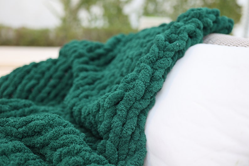 May include: A soft, green knitted throw blanket with a chunky cable knit pattern. The blanket is draped over a white surface.