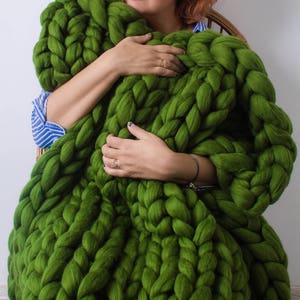 May include: A person is holding a large, chunky knit blanket made of thick green yarn. The blanket is very soft and fluffy, and it looks like it would be very comfortable to cuddle up in.