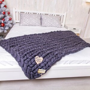 May include: A white bed with a gray chunky knit throw blanket. The bed has a white headboard and white bedding. The throw blanket is draped over the bed and has a heart-shaped tag attached to it.