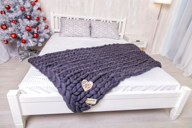 May include: A white bed with a gray chunky knit throw blanket. The bed has a white headboard and white bedding. The throw blanket is draped over the bed and has a heart-shaped tag attached to it.