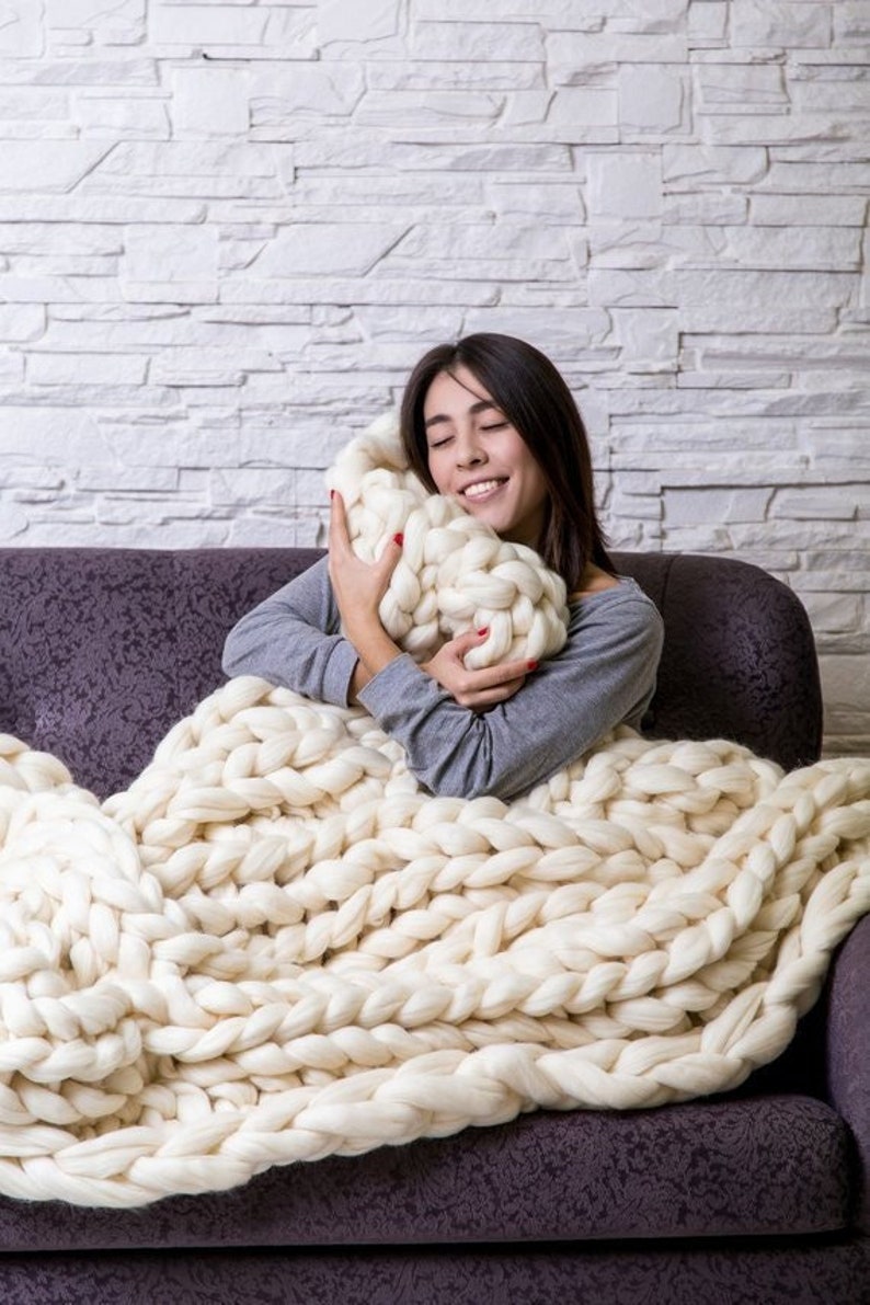 May include: A woman is sitting on a couch and hugging a large, chunky knit white blanket. The blanket is made of thick yarn and has a soft, cozy texture.