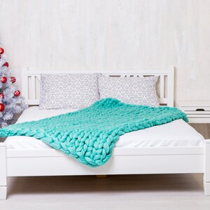 May include: A white bed frame with a white mattress and a teal knitted blanket. The bed is in a room with a white wall and a silver Christmas tree with red ornaments.