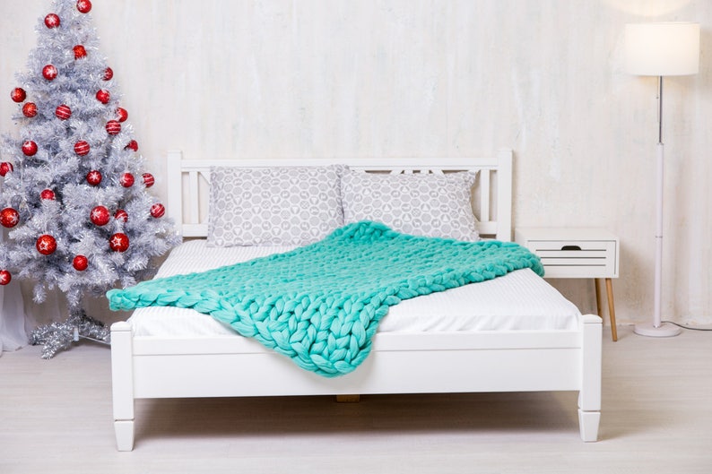 May include: A white bed frame with a white mattress and a teal knitted blanket. The bed is in a room with a white wall and a silver Christmas tree with red ornaments.