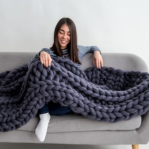 May include: A woman sits on a gray couch with a large, chunky knit gray throw blanket draped over her legs.