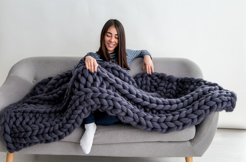 May include: A woman sits on a gray couch with a large, chunky knit gray throw blanket draped over her legs.