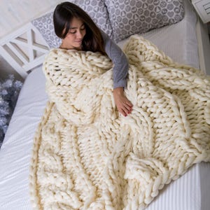 May include: A white chunky knit blanket with a cable knit pattern. The blanket is draped over a bed with white sheets and pillows.