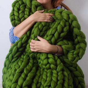 Op de afbeelding: Een groot, groen, grof gebreid deken gemaakt van dik garen. Het deken ligt op de schoot van een persoon en wordt vastgehouden door hun handen.