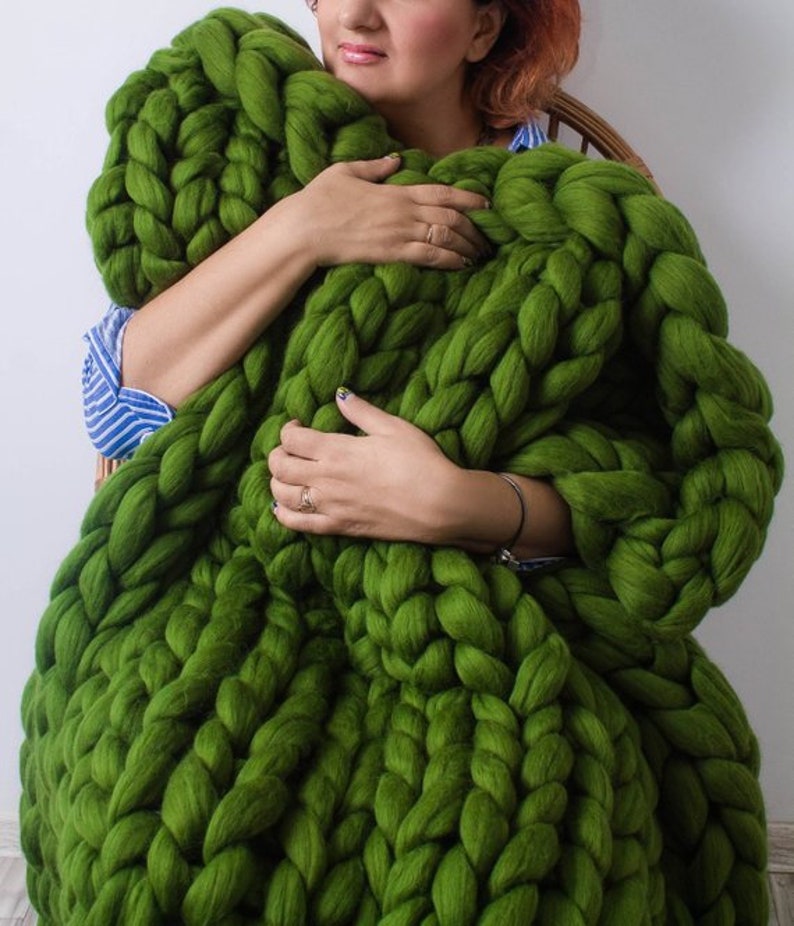 Op de afbeelding: Een groot, groen, grof gebreid deken gemaakt van dik garen. Het deken ligt op de schoot van een persoon en wordt vastgehouden door hun handen.
