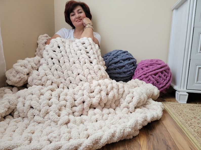 May include: A large, cream-colored chunky knit blanket with a woman sitting on it. There are two balls of yarn in the background, one is a dark blue color and the other is a light pink color.