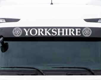 May include: A white truck cab featuring a black sun visor above the windshield. The visor displays the white text YORKSHIRE in capital letters, flanked on both sides by a circular white emblem depicting a stylized rose design.
