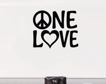 May include: A black vinyl decal applied to the white exterior of a camper trailer. The decal features the text ONE LOVE, with a peace symbol replacing the letter O in ONE and a heart shape replacing the letter O in LOVE, in a bold font.