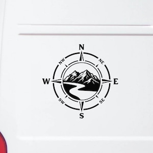May include: A black vinyl decal applied to a white vehicle surface, featuring a circular compass design with cardinal directions N, S, E, W and intercardinal points, surrounding a central graphic of mountain peaks and a winding river.