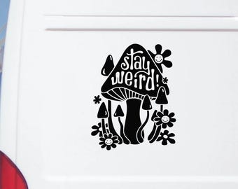 May include: A black vinyl decal applied to a white vehicle surface, featuring a large central mushroom with the text "stay weird!" written on its cap, surrounded by smaller mushrooms and several flowers with smiling faces in the center.