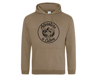 May include: A khaki-colored hooded sweatshirt featuring a black circular graphic on the chest. The design depicts mountains and pine trees with the text "Adventure is Calling" written in a cursive font inside the circle. The hoodie has long sleeves.