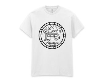 May include: A white short-sleeve t-shirt featuring a black circular graphic on the chest. The graphic depicts a camper van, a surfboard, ocean waves, and a sun, with the text Adventure Awaits written in a script font inside the circle.