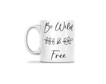May include: A white ceramic mug featuring the black printed text Be Wild and Free in a cursive script font. Between the words Wild and Free is a small black graphic of two leafy branches pointing outward from an ampersand symbol.