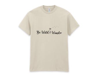 May include: A beige short-sleeve t-shirt featuring the black text Be Wild & Wander centered on the chest. The design includes a small dandelion graphic with seeds blowing in the wind positioned between the words Wild and Wander.