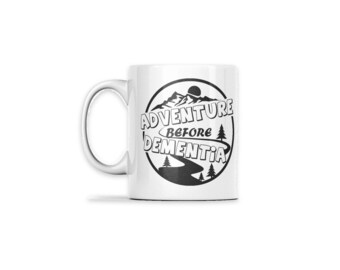 May include: A white ceramic mug featuring a black circular graphic. Inside the circle is a mountain landscape with pine trees and a winding road. The text reads ADVENTURE BEFORE DEMENTIA in bold, black, capital letters.