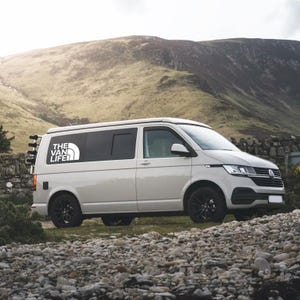 Puede incluir: Una furgoneta blanca con ventanas tintadas y una pegatina de "The Van Life" en el lateral. La furgoneta est&aacute; aparcada en una ladera rocosa con una monta&ntilde;a al fondo.