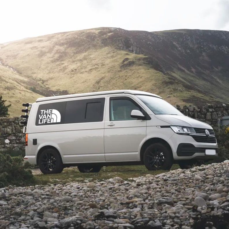Puede incluir: Una furgoneta blanca con ventanas tintadas y una pegatina de "The Van Life" en el lateral. La furgoneta est&aacute; aparcada en una ladera rocosa con una monta&ntilde;a al fondo.