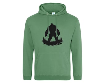 Puede incluir: Una sudadera con capucha de color verde salvia que presenta un estampado gráfico negro en el pecho. El diseño muestra la silueta de una criatura grande de pie en un entorno forestal con pinos y hierba en la base.