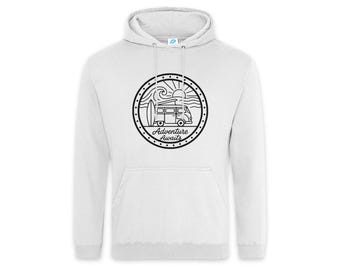 May include: A white hooded sweatshirt featuring a black circular graphic on the chest. The design depicts a van with a surfboard, waves, and a sun, accompanied by the text Adventure Awaits. The hoodie includes a front kangaroo pocket.