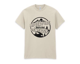 May include: A beige short-sleeve t-shirt featuring a black circular graphic on the chest. The design includes a mountain range, a winding road, pine trees, and the text ADVENTURE BEFORE DEMENTIA in a bold, stylized black font.