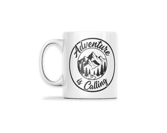 May include: A white ceramic mug featuring a black circular graphic. Inside the circle is a mountain range, pine trees, and a winding path. The text "Adventure is Calling" is printed in a black script font along the circular border.