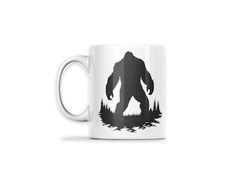 May include: A white ceramic mug featuring a black silhouette graphic of a large, hairy creature standing in a forest of pine trees. The design is centered on the front of the mug, which has a curved handle on the left side.