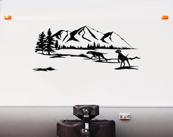 May include: A black vinyl decal applied to a white surface, featuring a silhouette of a mountain range, pine trees, and two dogs running through a landscape. The design is oriented horizontally and displays a detailed outdoor nature scene.