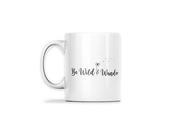 May include: A white ceramic mug featuring a black graphic of a dandelion with seeds blowing in the wind. The text "Be Wild & Wander" is printed in a black script font across the center of the mug, positioned next to the dandelion illustration.
