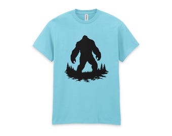 May include: A light blue short-sleeve t-shirt featuring a black graphic print of a large, hairy creature standing in a forest of pine trees. The design is centered on the chest area of the shirt, which has a crew neckline and a standard fit.
