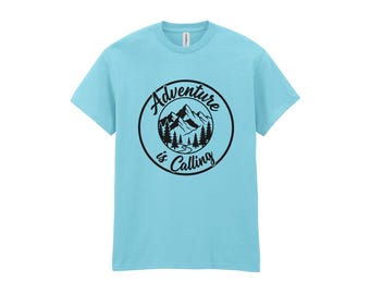 May include: A light blue short-sleeve t-shirt featuring a black circular graphic on the chest. The graphic depicts a mountain range and pine trees with the text Adventure is Calling written in a cursive font along the circular border.