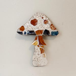 May include: A mosaic mushroom with a white stem and a speckled cap featuring orange and brown spots. The cap has a triangular shape with a dark blue border. The mushroom is a decorative item.