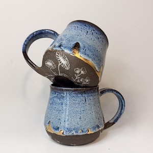May include: Two handmade ceramic mugs stacked on top of each other. The mugs have a blue speckled glaze with a dark brown base and handle. The bottom of the top mug and the top of the bottom mug have a gold accent. The top mug has a floral design.