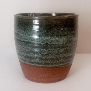 May include: A handmade ceramic cup with a dark teal and brown color scheme. The upper portion of the cup is a dark teal with a textured, layered appearance, while the bottom is a reddish-brown. The cup has a simple, cylindrical shape.