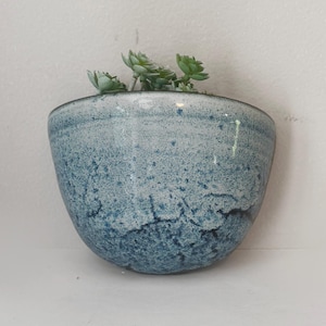 May include: A small, blue ceramic planter with a textured surface, holding a small succulent plant. The planter has a rounded shape and a speckled, mottled appearance, with shades of blue and white. The plant has green leaves.