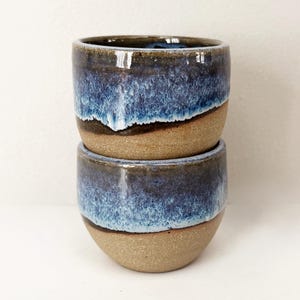 May include: Two stacked ceramic cups featuring a smooth, unglazed tan base and a reactive glaze top in shades of deep navy blue and lighter sky blue. The glaze drips down the sides, creating a textured transition between the blue and tan sections.