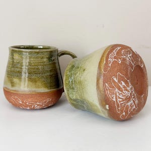 May include: Two handmade ceramic mugs. One is upright, the other is on its side. The mugs have a green and brown glaze with white etched designs on the base. The mugs are a unique addition to any kitchen.