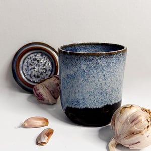 May include: A ceramic mug with a blue and black glaze, accompanied by a garlic bulb, cloves, and a small decorative plate. The mug has a cylindrical shape and a textured surface. The plate has a brown and blue rim.
