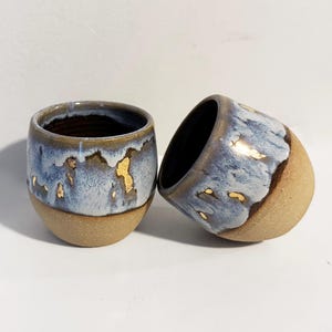 May include: Two small ceramic cups with a rounded shape. The lower portion features a textured, matte beige finish, while the upper section is glazed in a mottled blue and white pattern with small, irregular gold-coloured accents throughout the glaze.