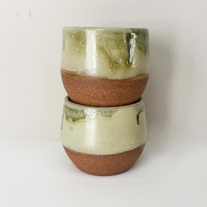 May include: Two handmade ceramic cups stacked on top of each other. The cups have a rounded shape with a reddish-brown base and a light green and cream-colored upper section. The glaze has a dripped effect.