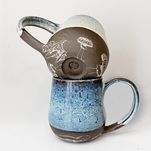 May include: Two ceramic mugs stacked on top of each other. The bottom mug is blue with a speckled glaze, and the top mug is brown with white mushroom illustrations. Both mugs have handles and a unique, handmade appearance.