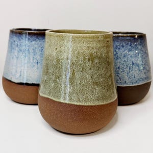 May include: Three ceramic stemless wine glasses. The glasses have a rounded base and a slightly tapered top. The bottom half of each glass is a reddish-brown color, while the top half is glazed in shades of blue and green.