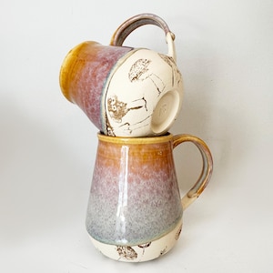 May include: Two stacked ceramic mugs featuring a gradient glaze that transitions from a warm golden-yellow rim to a soft mauve and grey base. The lower portion of each mug is cream-colored and decorated with brown line drawings of various mushrooms.