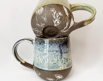 May include: Two handcrafted ceramic mugs. The top mug has a tan upper section and a dark brown lower section with white floral designs. The bottom mug is blue and brown. Both mugs have handles and are stacked.